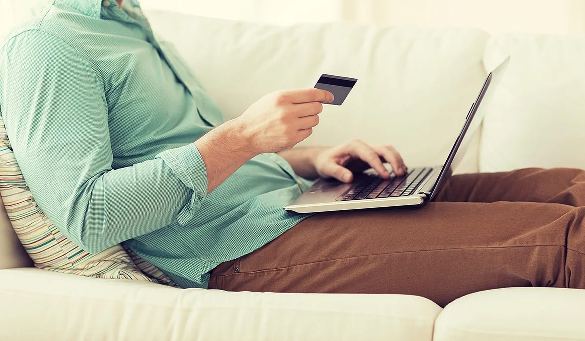 Person using a laptop and holding a credit card on a cozy couch.