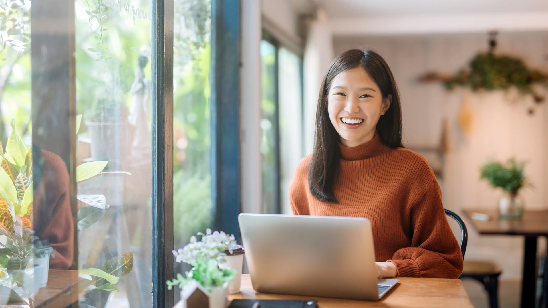 Smiling woman in cozy cafe working on a laptop near a window with plants.